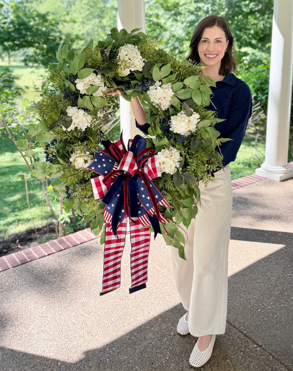 Woman holding a large decorative wreath with an American flag bow on a porch.