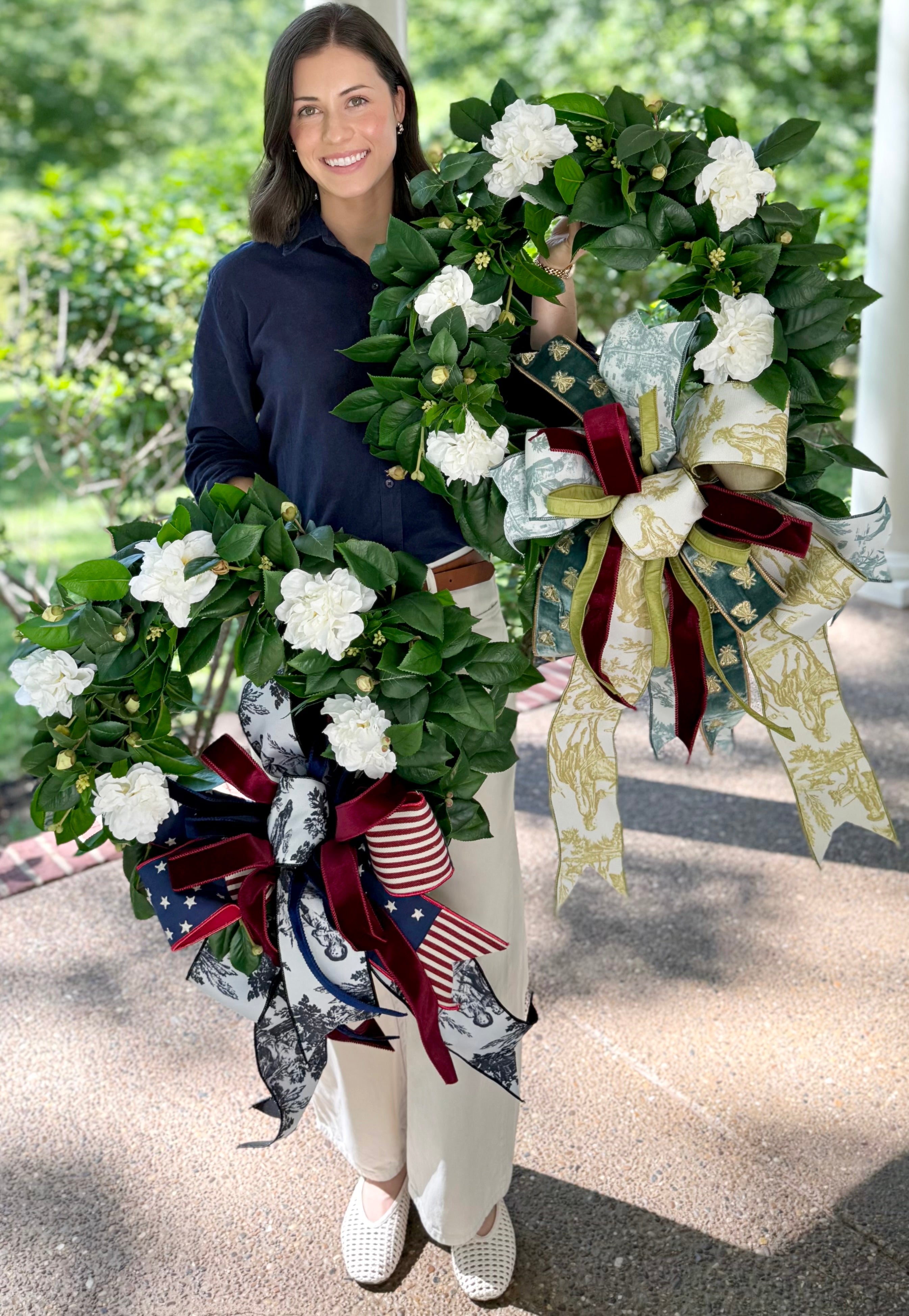 Woman holding two decorative wreaths with flowers and ribbons outdoors.